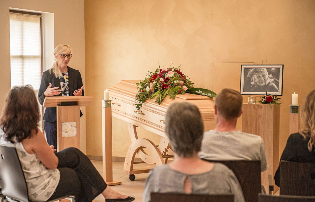 Liebevoll gestaltet Trauerfeier bei Kuhler & Kaufmann Bestattungen mit Trauerrednerin neben Sarg mit Dekoration und Trauergemeinde